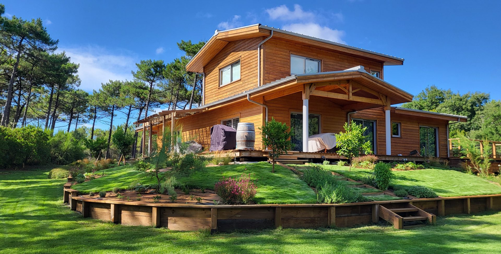 Maison en bois entourée de pins, avec un jardin en terrasses soutenues par des rondins et des plantations méditerranéennes
